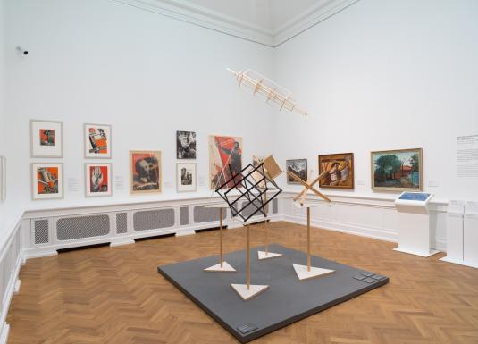 Izstādes zāle ar 20. gadsimta mākslas darbiem — gleznām, fotomontāžām un konstruktīvisma iedvesmotām telpiskām instalācijām, eksponētām uz baltām sienām un koka grīdas fona.