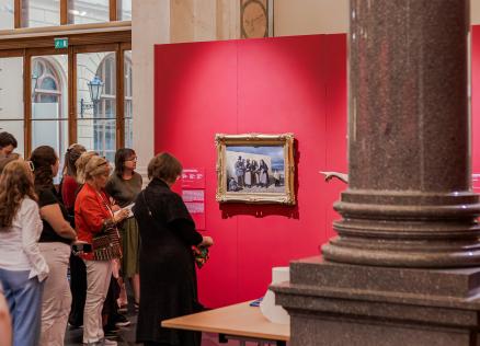 Apmeklētāji aplūko gleznu uz spilgti sarkanas sienas muzeja izstāžu zālē.