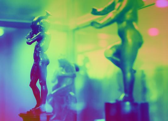 Mazas bronzas figūriņas ar cilvēku tēliem stiklotā vitrīnā krāsainā apgaismojumā.