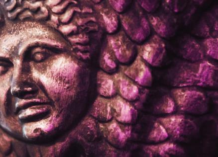 Attēls ar violetu toni pārklātu antīka stila bareljefa fragmentu – tuvplāns ar mitoloģiska tēla seju un zvīņainām detaļām fonā.