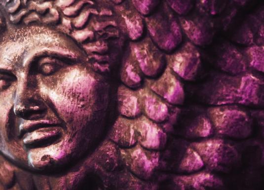 Attēls ar violetu toni pārklātu antīka stila bareljefa fragmentu – tuvplāns ar mitoloģiska tēla seju un zvīņainām detaļām fonā.