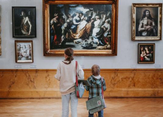 Divi bērni ar pleca somām muzeja zālē aplūko gleznas ar zeltītiem rāmjiem.