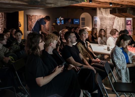 Auditorija klausās lekciju izstāžu telpā ar koka sijām un izstādītiem darbiem fonā.