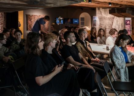 Auditorija klausās lekciju izstāžu telpā ar koka sijām un izstādītiem darbiem fonā.