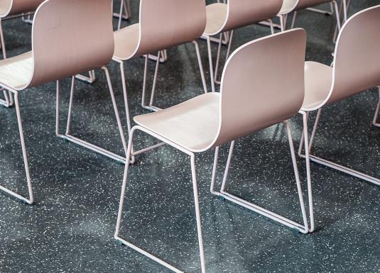 Tuvplāns ar gaišiem krēsliem, kas novietoti rindās uz tumša, plankumaina grīdas seguma.