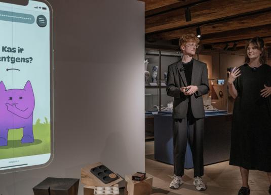 Prezentācija izstādē – divi cilvēki stāv pie ekrāna, kur projicēts mobilās lietotnes attēls ar jautājumu “Kas ir rentgens?”.