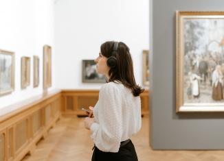 Jauna sieviete klausās audiogidu un aplūko gleznas Latvijas Nacionālā mākslas muzeja zālē.