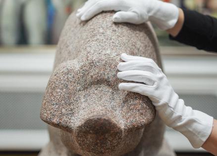 Rokas baltos cimdos pieskaras granīta skulptūrai muzeja ekspozīcijā.