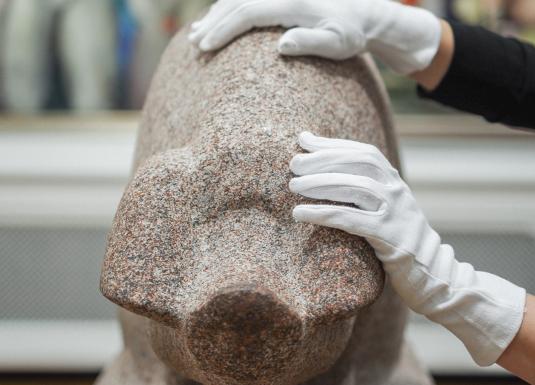 Rokas baltos cimdos pieskaras granīta skulptūrai muzeja ekspozīcijā.