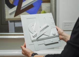 Persona tur rokās taustes kopiju ar reljefu, kas veidota pēc kubisma stila gleznas motīviem, ļaujot mākslu izjust ar pieskārienu.