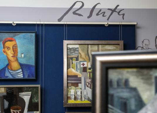 Gleznu ekspozīcija ar cilvēku portretiem un pilsētas ainavu zilā fonā.