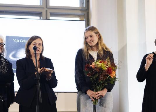 Četras sievietes stāv uz skatuves Latvijas Nacionālajā mākslas muzejā Vijas Celmiņas fonda stipendijas pasākumā, viena no viņām tur puķu pušķi.