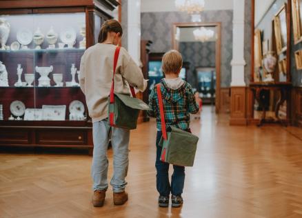 Divi bērni ar plecu somām dodas cauri muzeja zālei, kurā izstādīti porcelāna trauki un gleznas klasiskā interjerā.